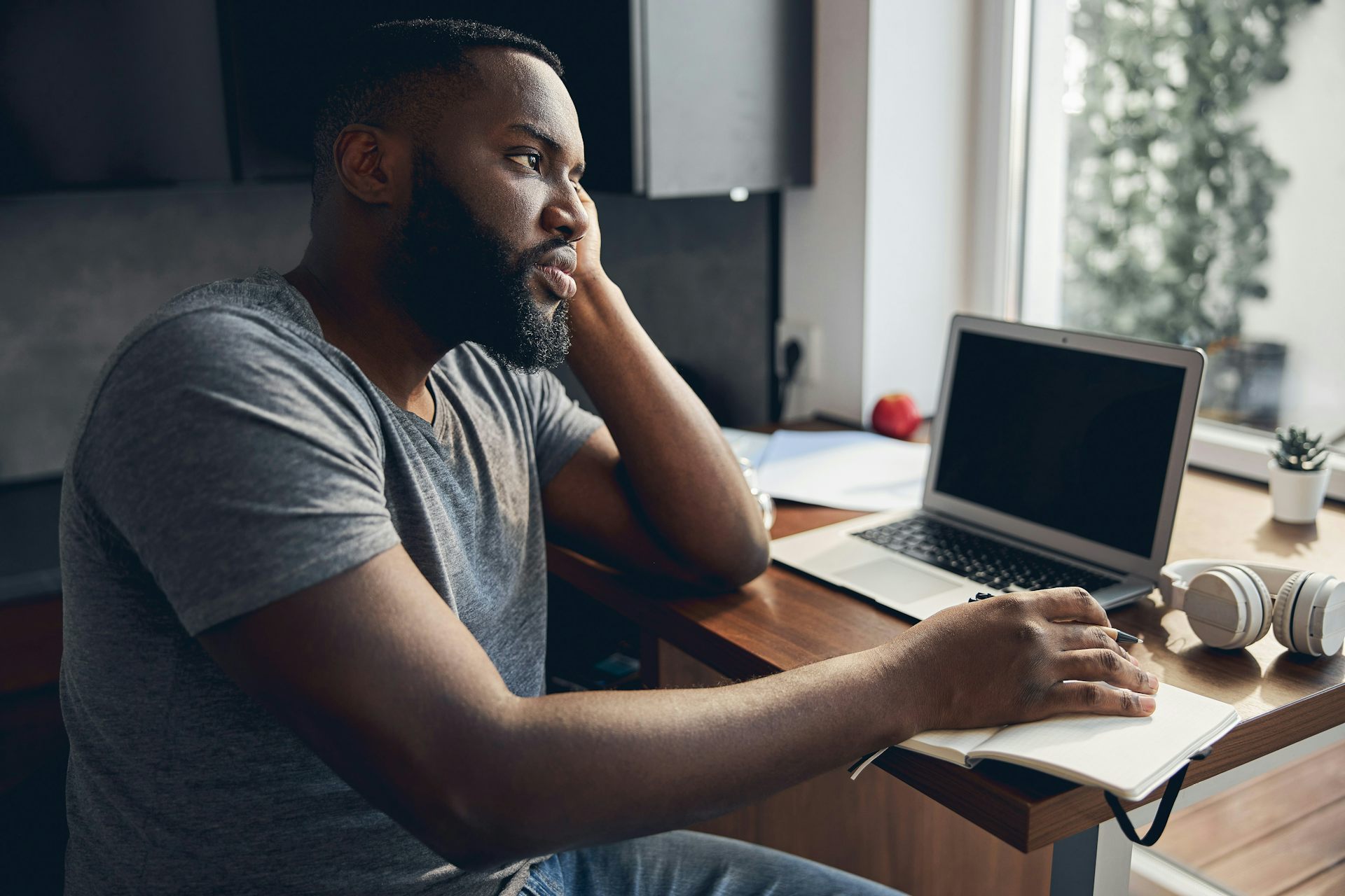 Un homme est assis &agrave; un bureau, un ordinateur portable ouvert devant lui, le regard tourn&eacute; vers la fen&ecirc;tre. 