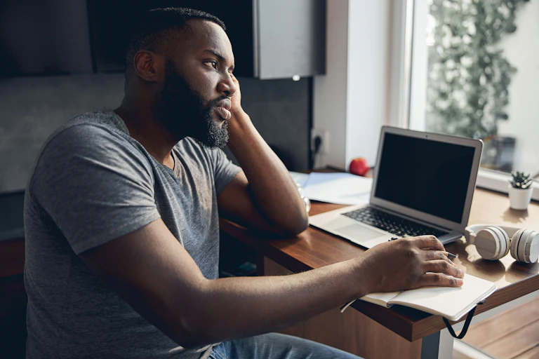 Un homme est assis à un bureau, un ordinateur portable ouvert devant lui, le regard tourné vers la fenêtre.