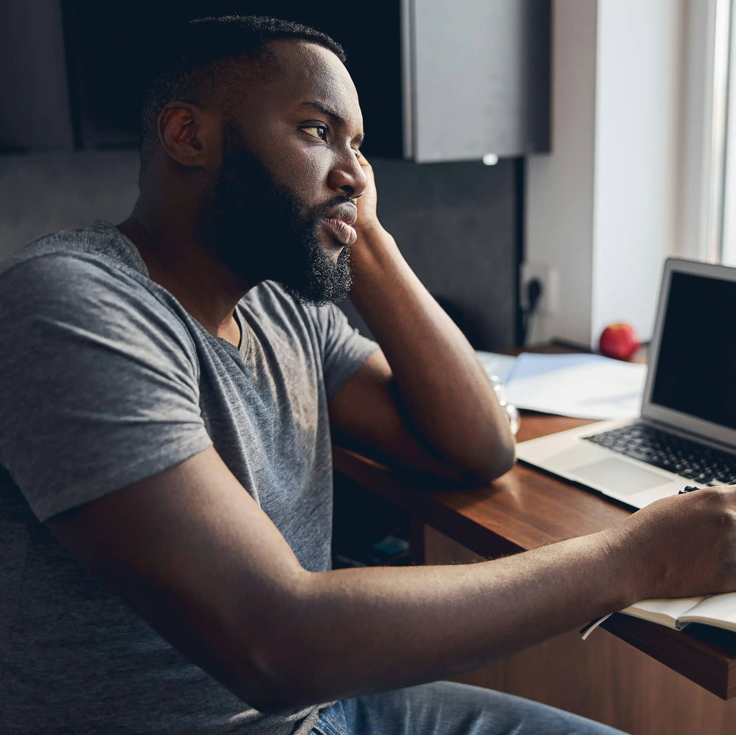 Un homme est assis à un bureau, un ordinateur portable ouvert devant lui, le regard tourné vers la fenêtre.