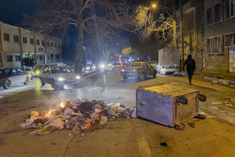 A dumpster on its side is on a street with cars.