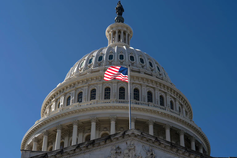 An American flag flies in the wind in front of a white domed building