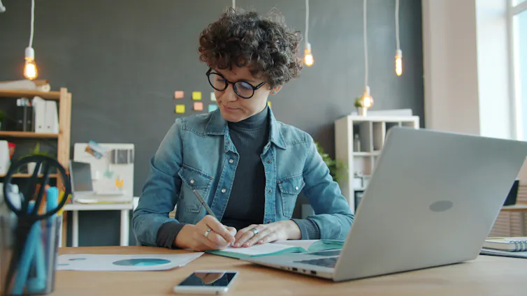 Woman at work on laptop writing in notebook