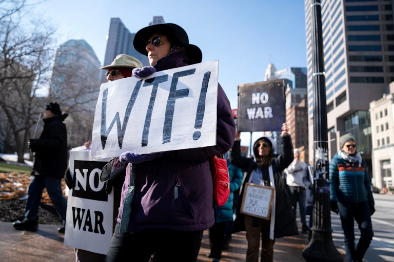 A protester holds a sign that reads WTF. Another person behind him holds a sign that reads No War.