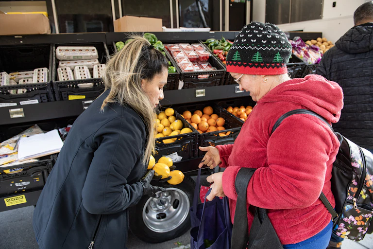 One person helps another select fruit from a display of various foods.