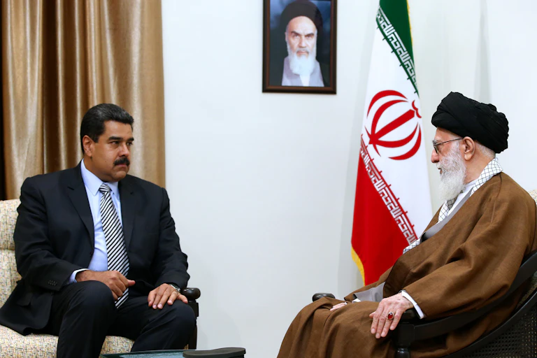 Two men, one in a dark suit and the other in a flowing brown robe, sit and talk to each other.
