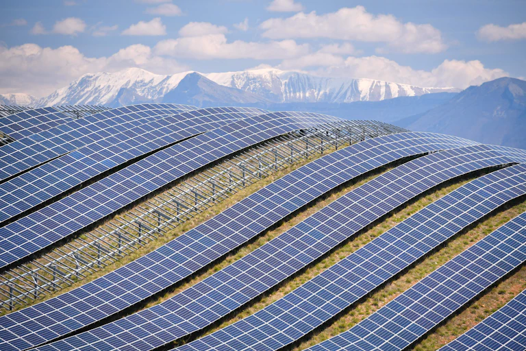 Vista panorâmica das faixas de painéis solares numa encosta com montanhas cobertas de neve ao fundo.
