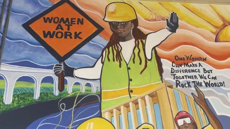 A mural of a women in a construction hard hat holding a sign that says 'women at work'