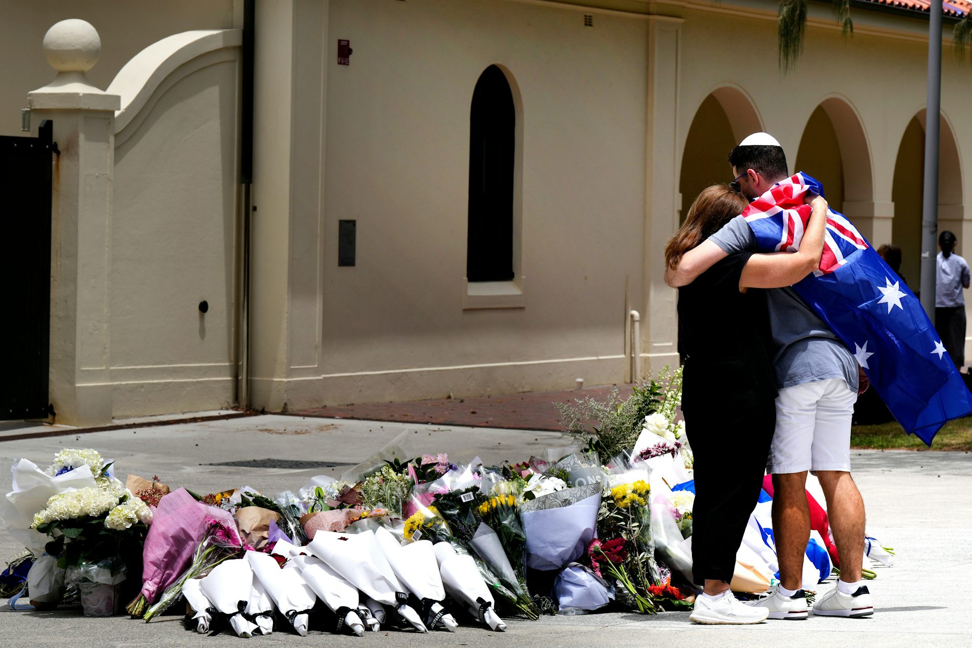 A woman hugs a Jewish man wearing an Australian flag in front of a memorial of flowers