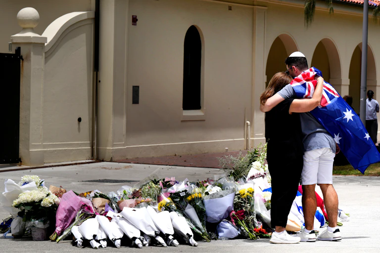 A woman hugs a Jewish man wearing an Australian flag in front of a memorial of flowers