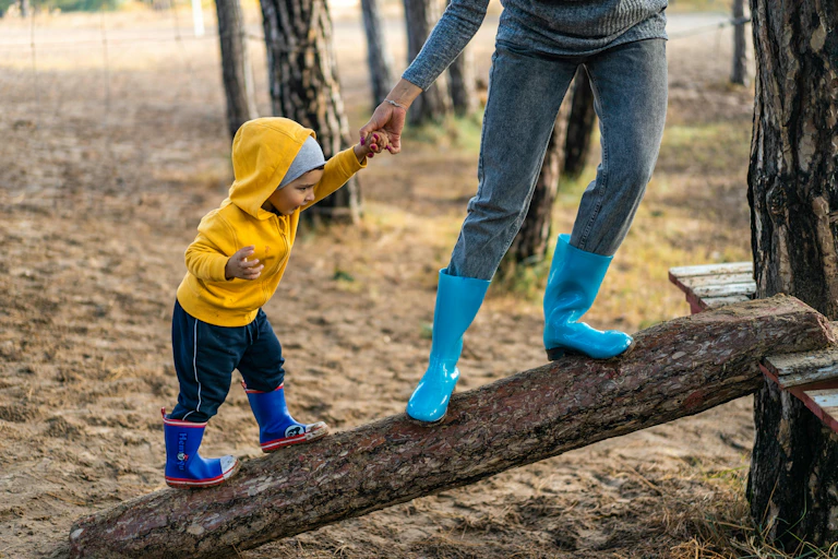 A parent in blue gumboots helping a child in yellow jumper climb over a tree trunk.