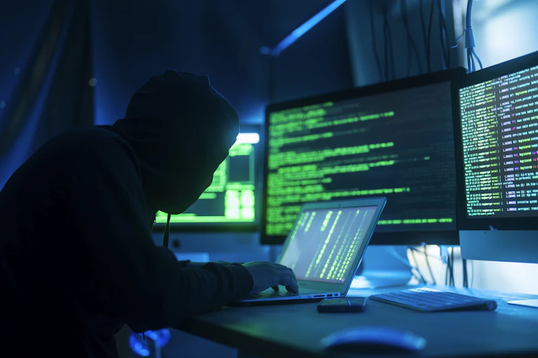 Shadowy figure of a person using a computer, with computer screens in the background
