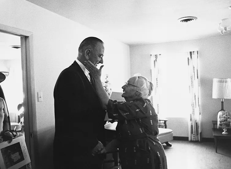 An older woman pats the cheek of a much taller middle-aged man.