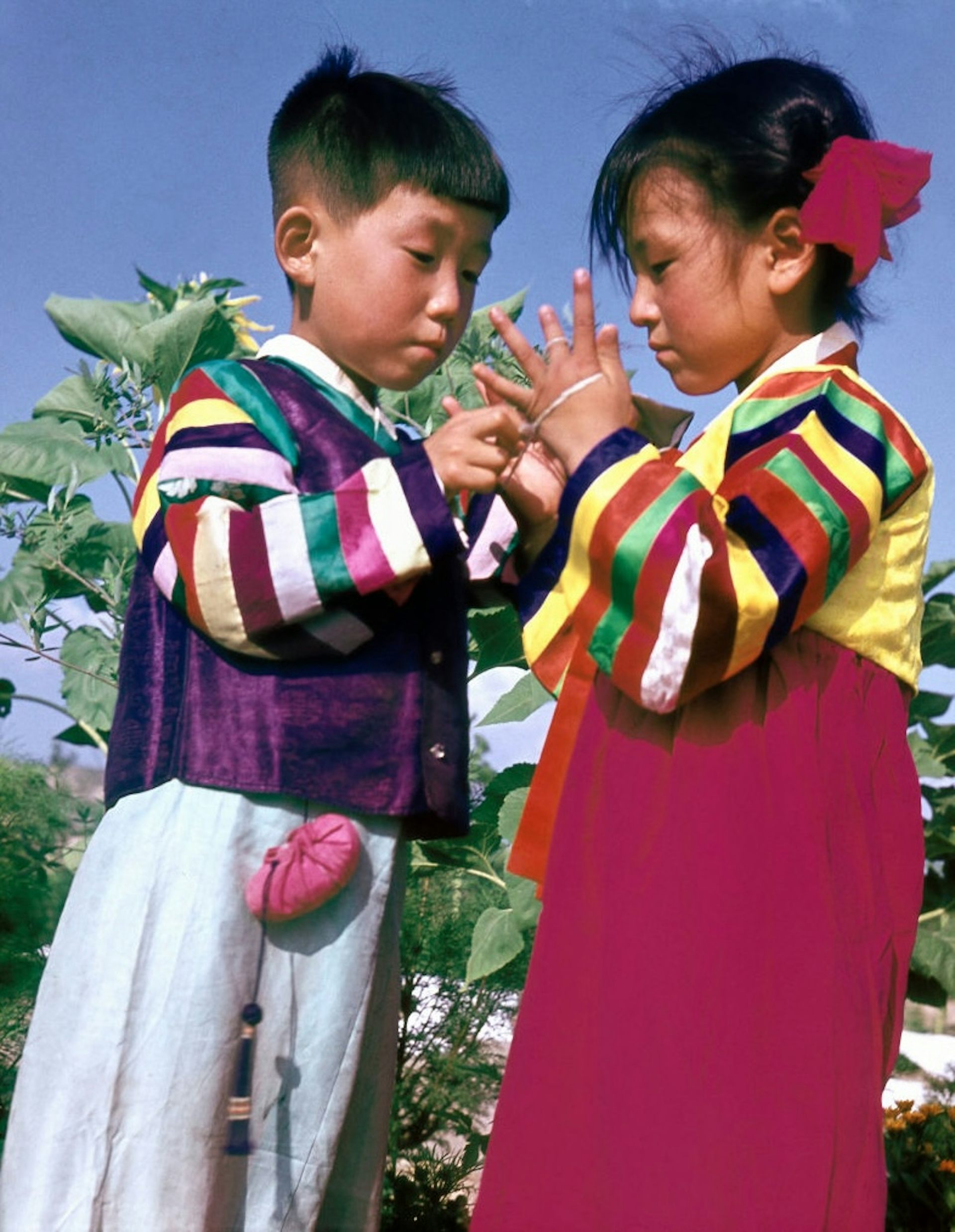Two young Asian children wearing colorful outfits playing a game using a string wound through their hands.