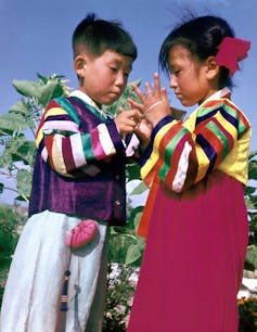 Two young Asian children wearing colorful outfits playing a game using a string wound through their hands.