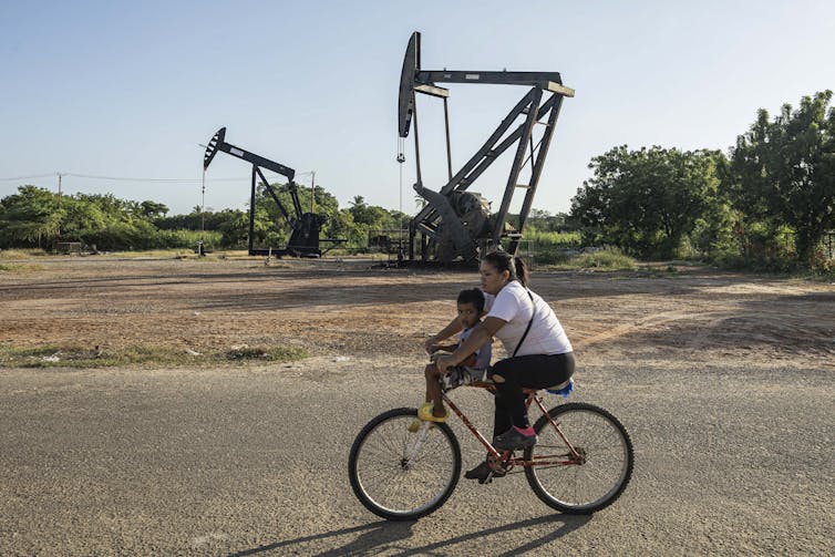a woman and child cycle past a pair of oil derricks in venezuela