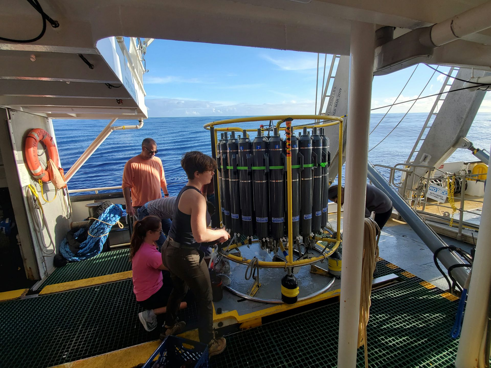 Scientists aboard a research vessel prepare a large device with many tubes for collecting samples once lowered into the ocean.
