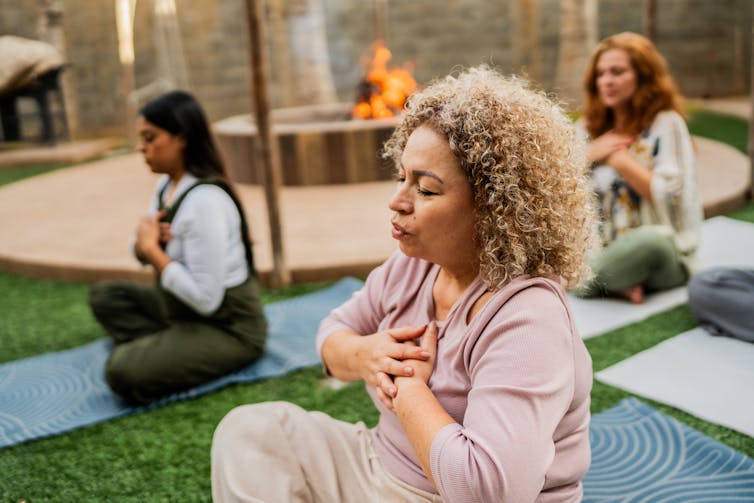 Three women sit on yoga mats, eyes closed, palms resting over their hearts in meditation.