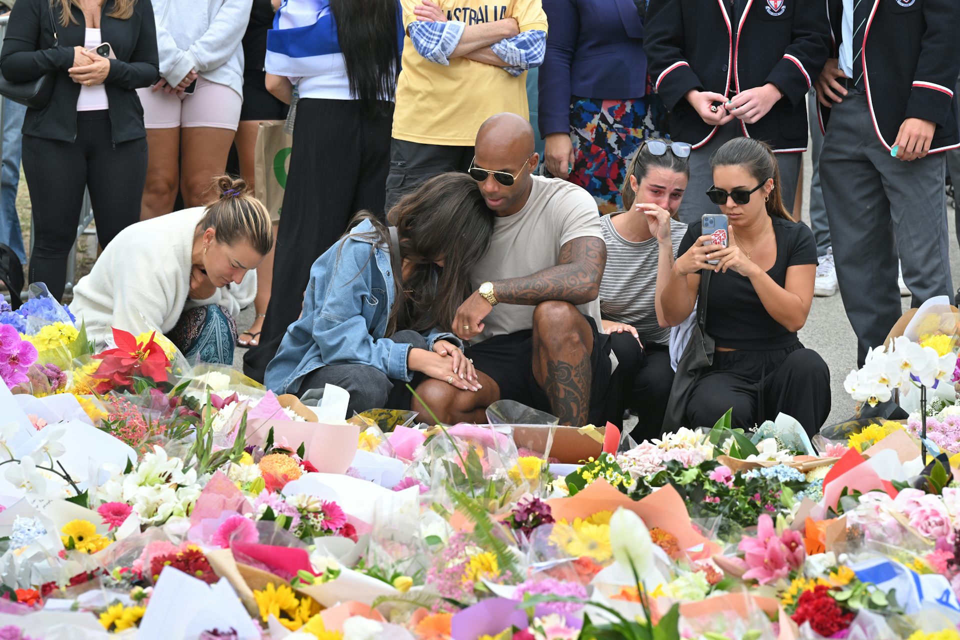 A group of mourners kneel beside an array of flowers at a memorial