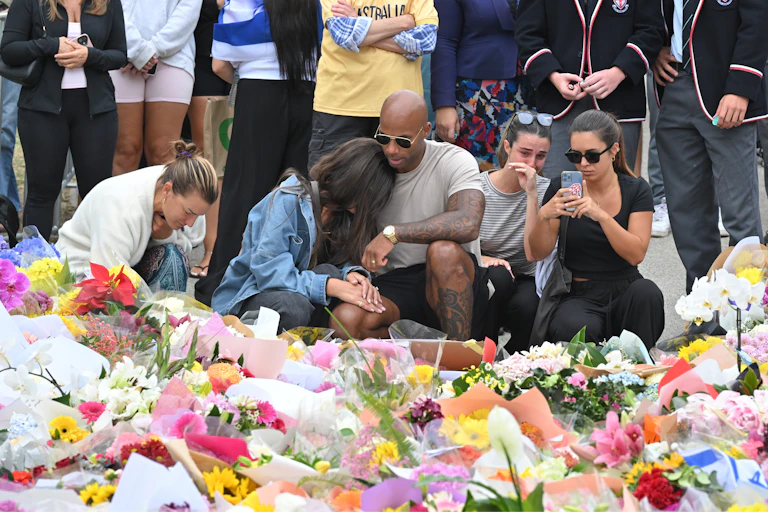A group of mourners kneel beside an array of flowers at a memorial