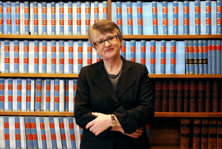 A woman with glasses stands in front of a large bookcase