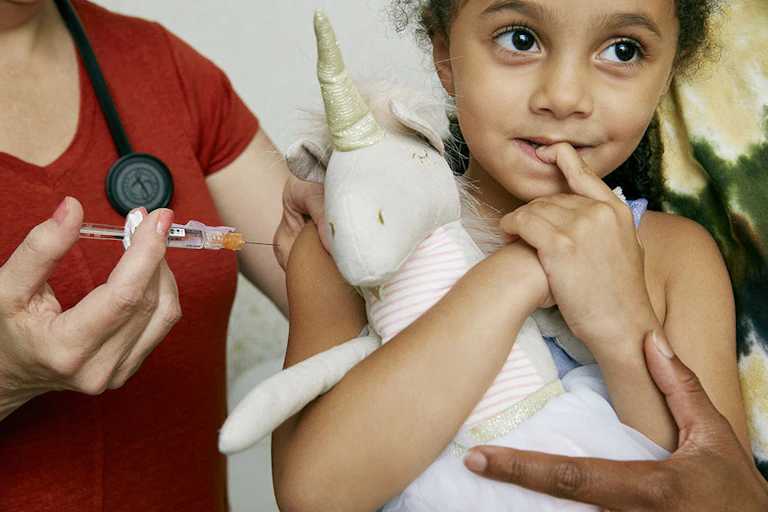 A health care worker gives a vaccine to a young child holding a stuffed unicorn.