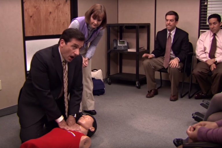Person in suit kneeling with hands on a CPR mannequin, other people looking on