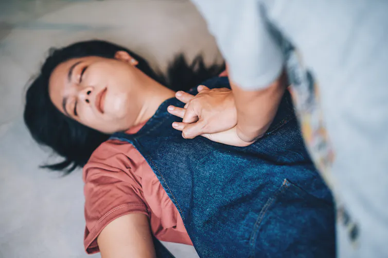 Woman lying unconscious on ground, another person's interlaced hands on her chest