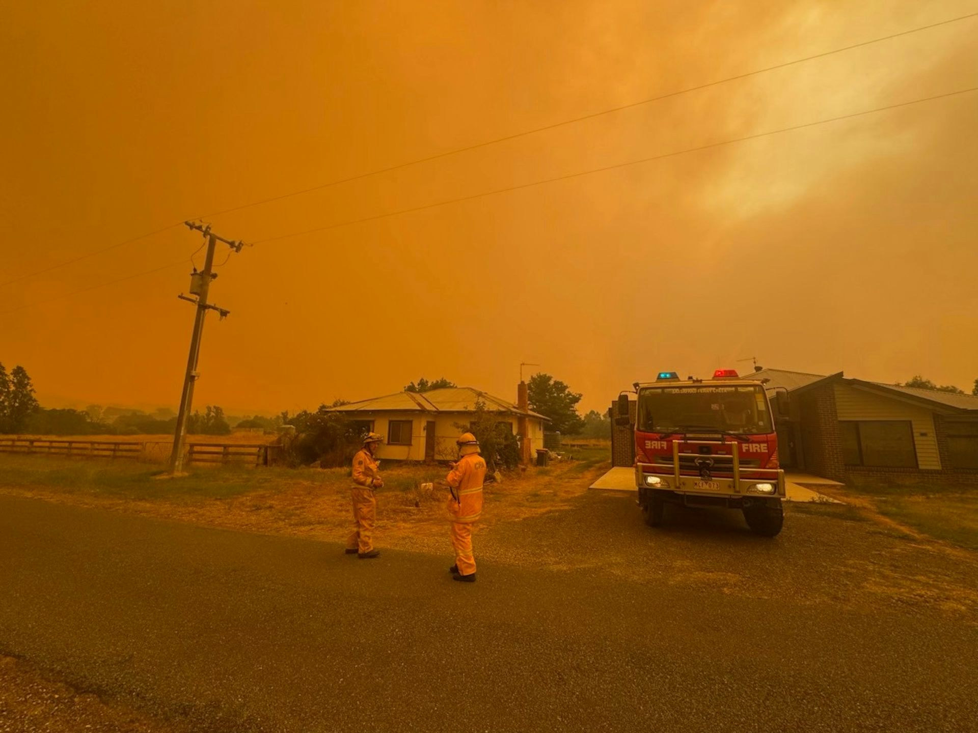 Two firefighters stand in front of a house with a yellow, smoky sky behind them.