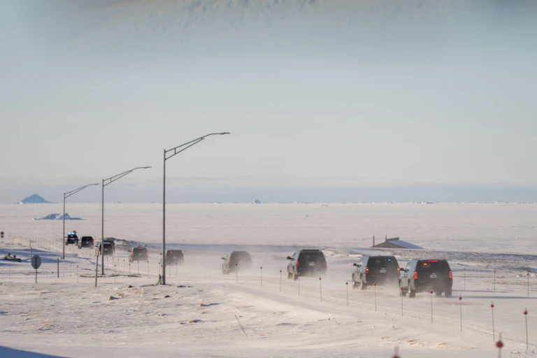 A line of vehicles drives across a long straight road in the Arctic.