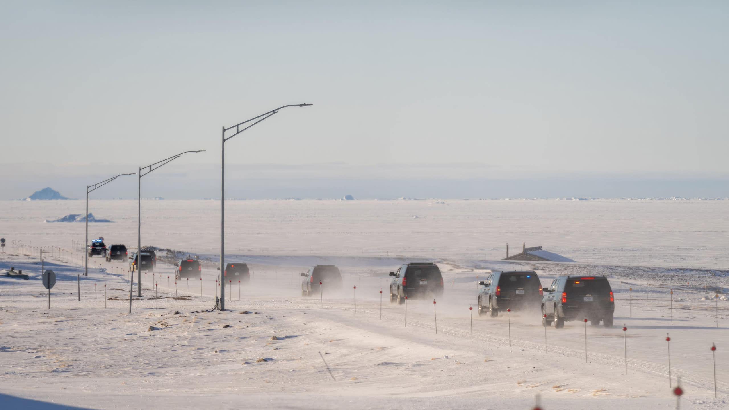 A line of vehicles drives across a long straight road in the Arctic.