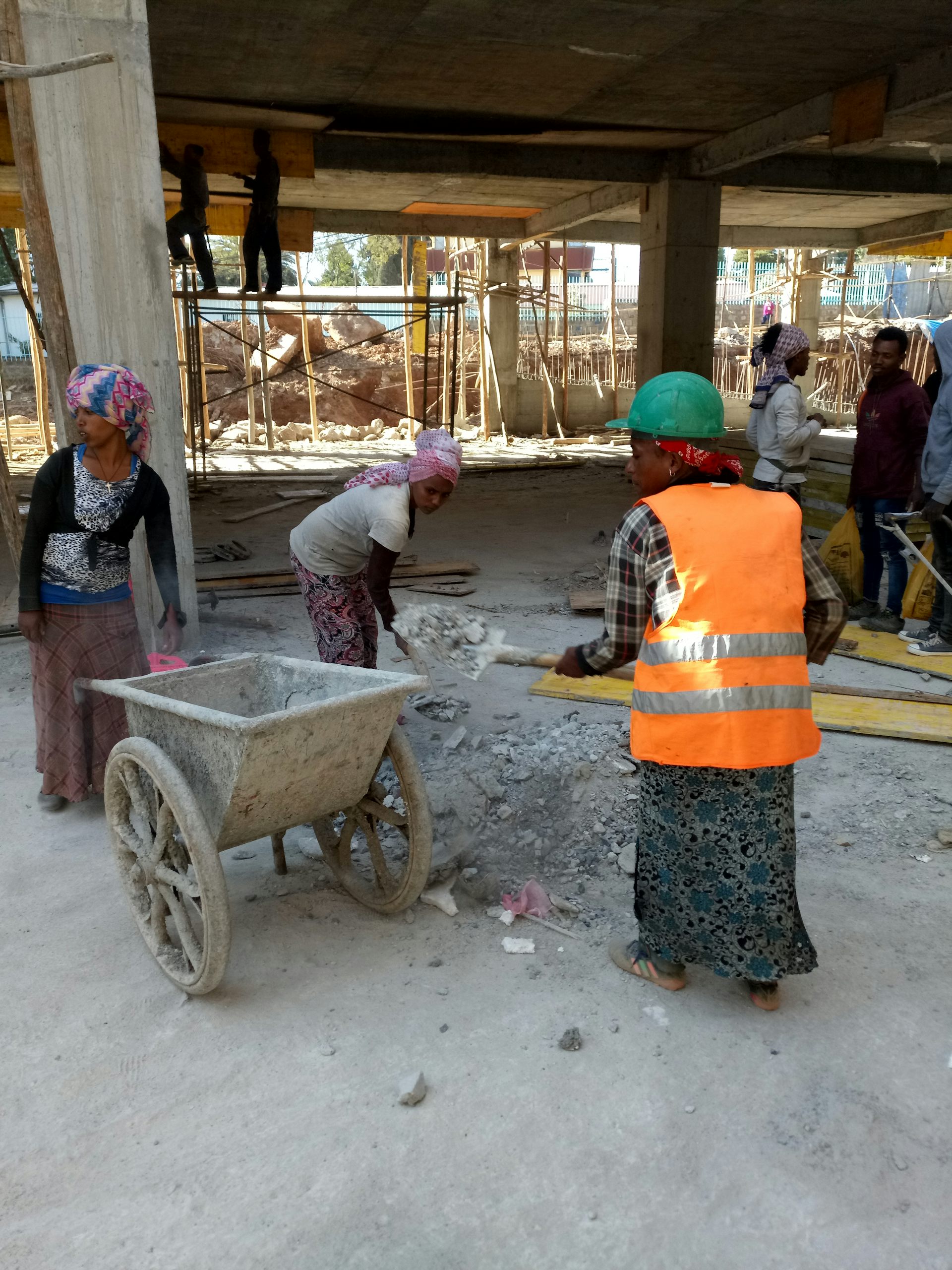 A woman in a high-visibility jacket loading debris into a wheelbarrow at a construction site as other women help