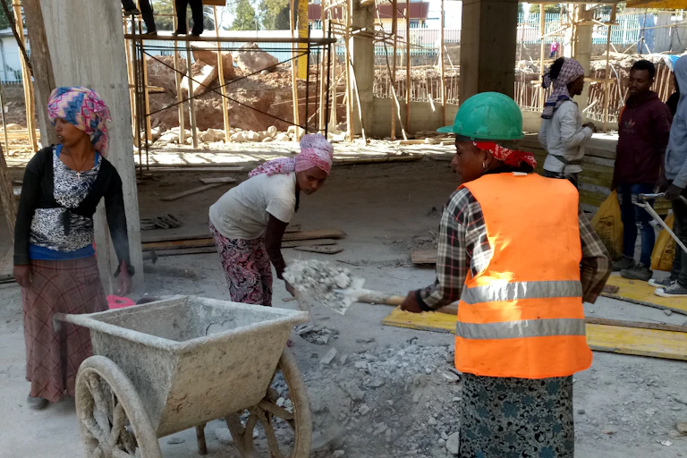 A woman in a high-visibility jacket loading debris into a wheelbarrow at a construction site as other women help