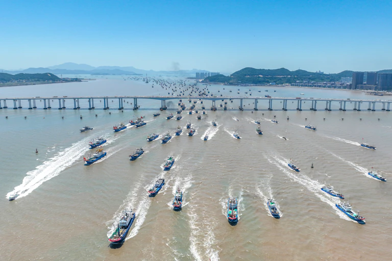 A fleet of dozens of fishing boats streams under a bridge.
