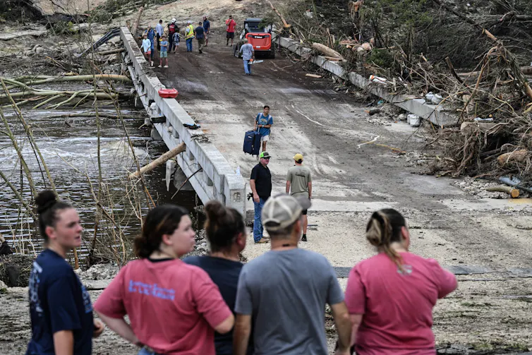 People stand near a bridge and searchers look through debris that has washed up.