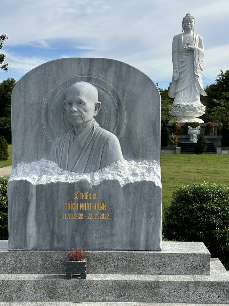 A monk’s gravestone rests in the foreground, with a towering statue of the Buddha behind it.