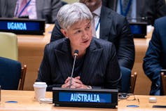A woman with short grey hair sits at a desk behind an Australia sign.