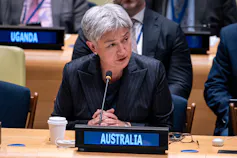 A woman with short grey hair sits at a desk behind an Australia sign.
