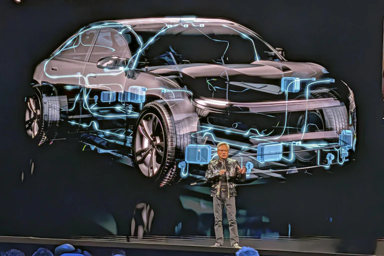 Nvidia's CEO Jensen Huang on stage in a shiny leather jacket, standing in front of a futuristic image of a car.