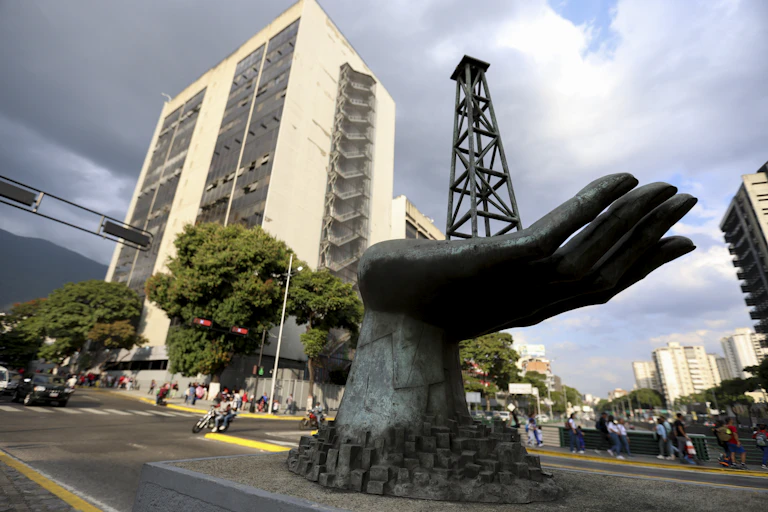 A large metal hand holding an oil drill rig sits in a plaza in front of a tall office building.