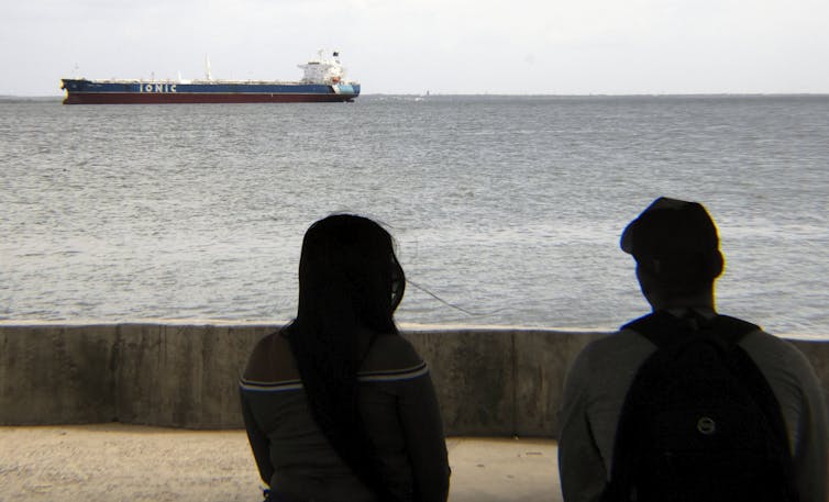 Why unlocking Venezuelan oil received’t imply a lot for US power costs 1 Two figures watch a large ship move across the water.