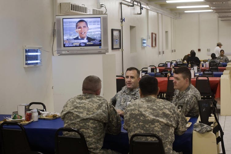 US soldiers sitting at a table with a tv behind them showing an image of Barack Obama.