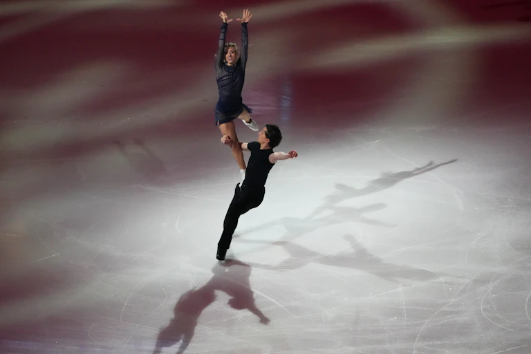 A figure skater lifts another on his hip on the ice