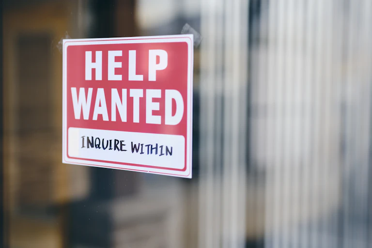 A red sign with white writing says 'Help wanted, inquire within' on a glass door.