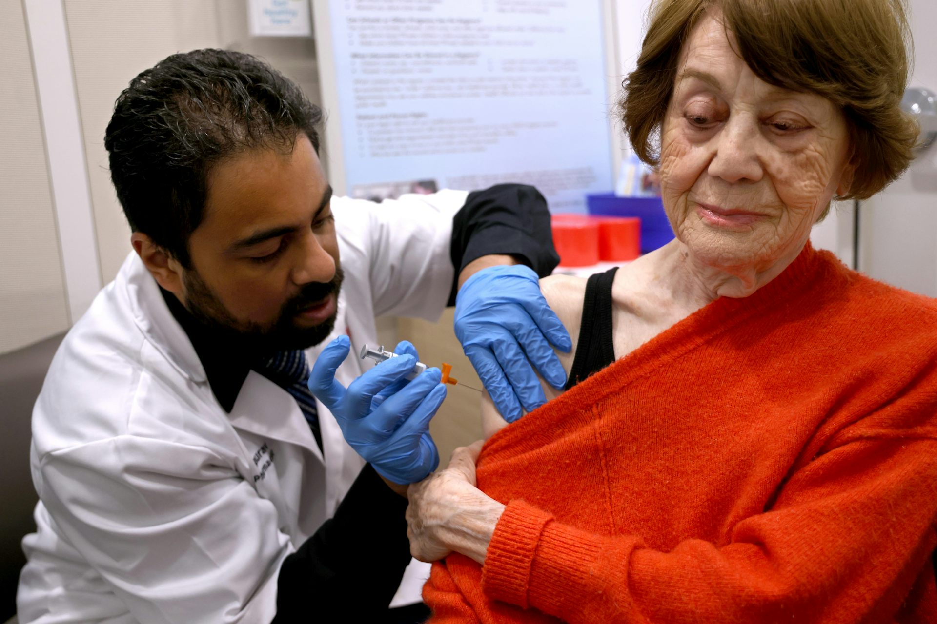 An older woman in a red sweater is getting a shot in her arm by a man in a doctor's coat with blue gloves on.