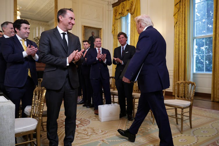 A man in a blue suit walking into a group of other men in suits in a high-ceilinged room.