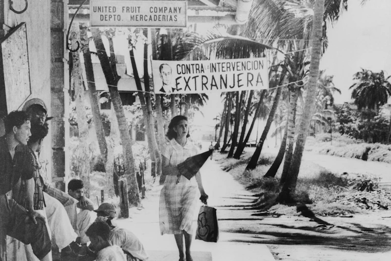 A woman walk down a palm tree lined sidewalk, past signs saying 'United Fruit Co. Depto de Mercaderias' and 'Contra intervencion extranjera.'