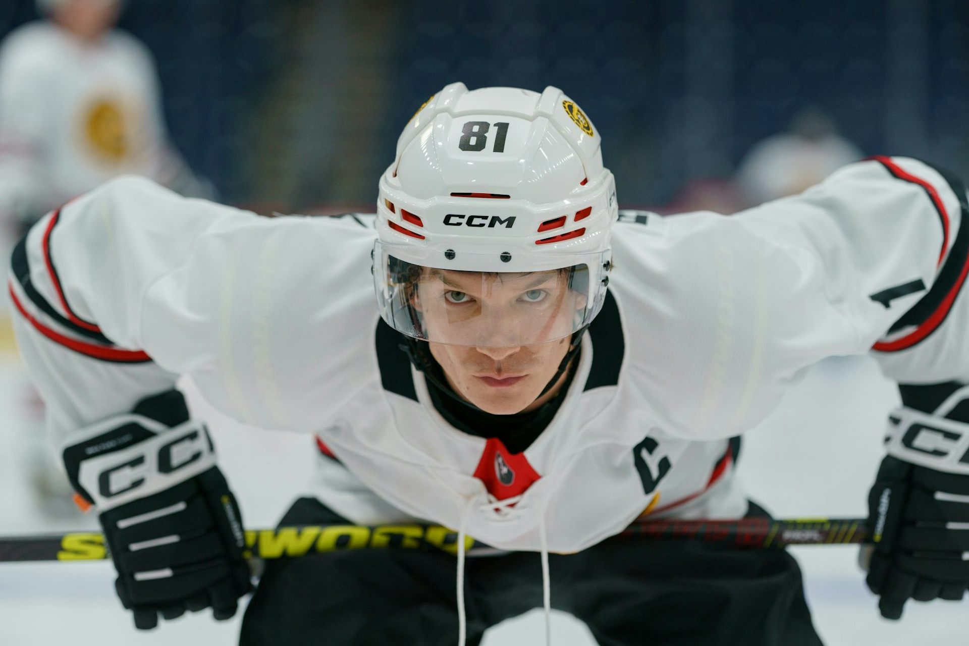 A white man in a hockey uniform leans over while holding his stick against his thighs