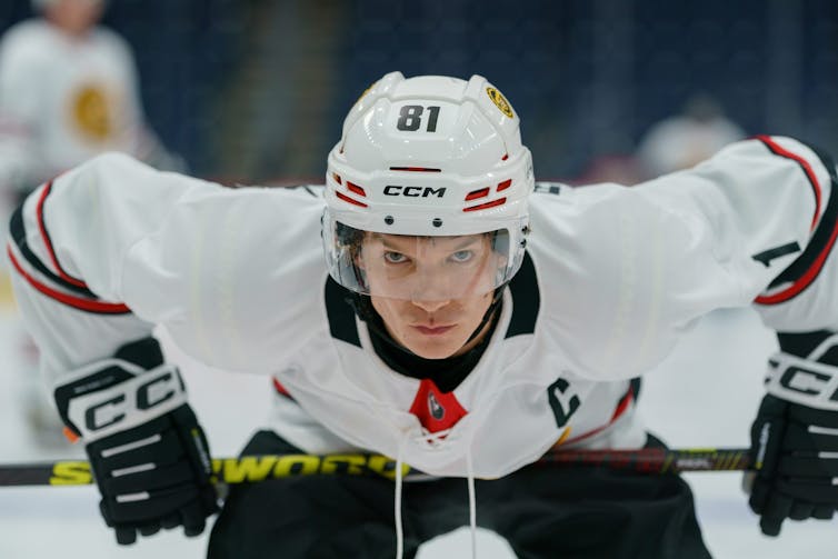 A white man in a hockey uniform leans over while holding his stick against his thighs