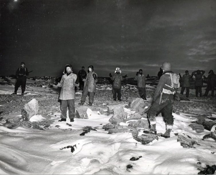 Men holds their hands in the air in surrender while soldiers point guns at them.