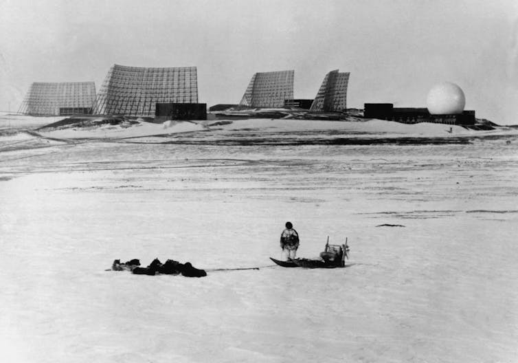A person stands next to a sled and dog team looking at a large radar installation.